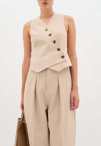 Top beige senza maniche con scollo a V e orlo asimmetrico, caratterizzato da sei bottoni scuri. Abbinato a pantaloni beige plissettati a vita alta.