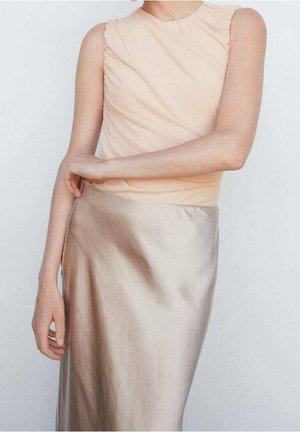 Personne portant un haut beige sans manches et une jupe en satin beige clair brillant, avec un bras plié et l'autre détendu le long du corps.