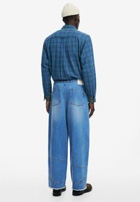 Camisa de cuadros azul de manga larga y jeans de mezclilla azul claro holgados. Los jeans tienen costuras visibles y dos bolsillos traseros. Diseñados con un ajuste relajado.