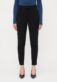Pantalones negros ajustados de tela suave, con una cintura elástica ajustable, bolsillos laterales y una textura lisa. Llevados con tacones negros.
