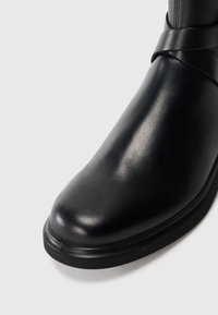 Black leather ankle boot with a rounded toe, smooth texture, and a wide strap accent around the upper, featuring a flat rubber sole.