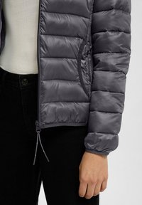 Grijze, gewatteerde jas met een gladde, glanzende textuur. Heeft elastische cuffs, zijzakken en een ritsluiting met een trekkoord aan de onderkant.