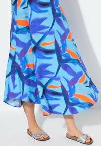 Floral Maxi-Kleid in Blau mit orangefarbenen und lila abstrakten Mustern. Model trägt graue Rutsch-Sandalen mit Korksohle.