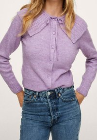 Donna che indossa un cardigan button-down di un leggero viola con un ampio colletto e jeans in denim blu a vita alta, con le mani nelle tasche.