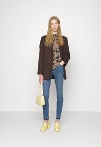 Blazer oversized castanho por cima de uma blusa floral, jeans skinny azuis, sapatos de salto bloco amarelos e uma pequena bolsa amarela.