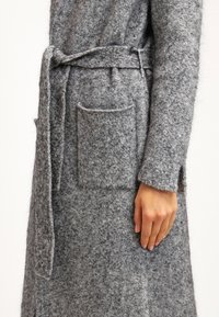 Manteau en laine gris texturé avec une ceinture en tissu nouée à la taille et une grande poche appliquée sur le côté, porté par une personne.