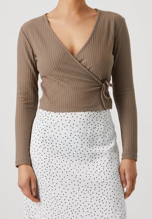 Femme portant un haut cache-cœur côtelé marron à manches longues et une jupe blanche à pois noirs sur un fond uni.