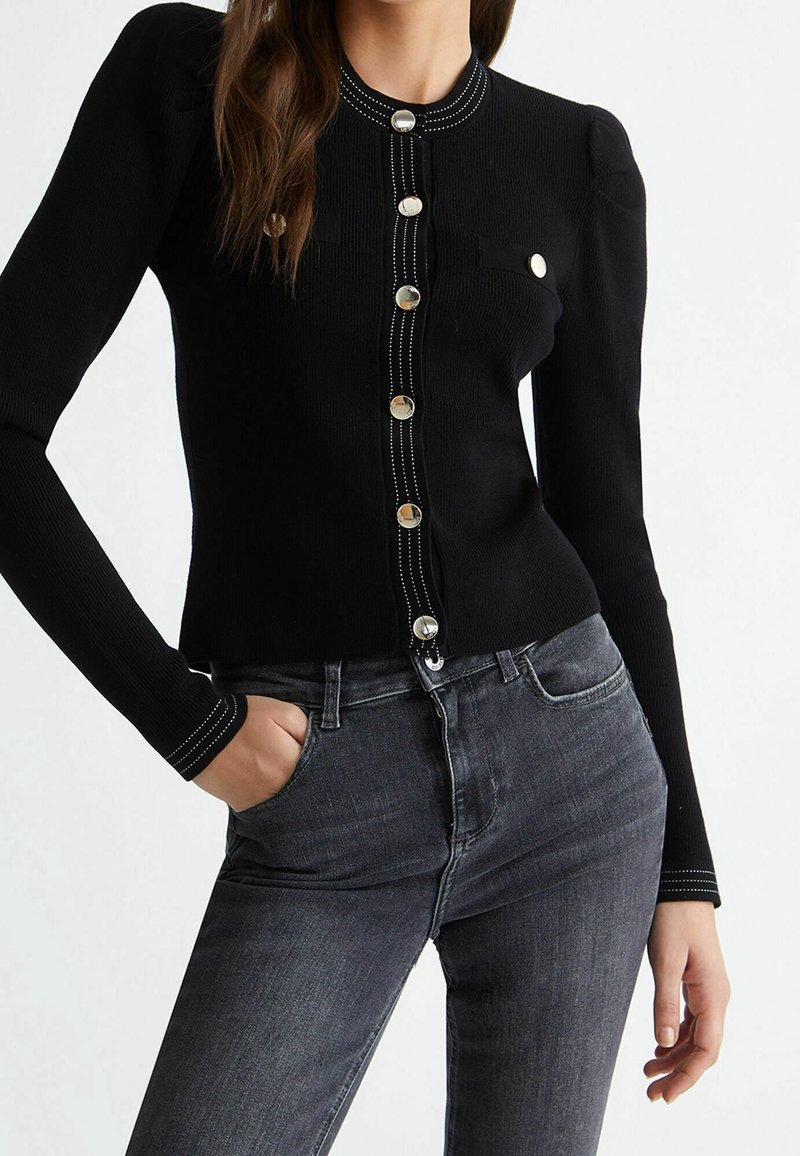 Sort ribstrikket cardigan med rund krave og sølvknapper, har kontrasterende hvide syninger og lange ærmer, kombineret med mørkegrå jeans.