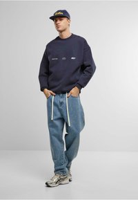 Hanorac naval cu logo-uri frontale, pantaloni relaxați din denim albastru și adidași argintii, combinați cu o șapcă închisă la culoare cu text galben.