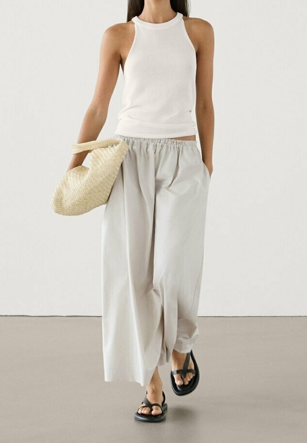 Femme portant un haut blanc sans manches, un pantalon ample gris clair, des sandales noires, tenant un sac en paille tressée, marchant sur un sol gris uni.