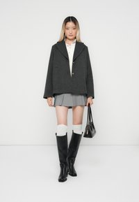 Veste en laine grise mélangée avec un col châle, associée à une jupe plissée grise, des bottes noires jusqu'aux genoux et un petit sac à main noir.