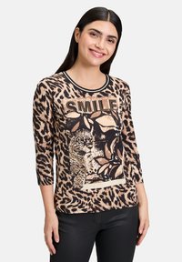 Haut à manches longues imprimé léopard avec des bordures noires, affichant un graphique de léopard et le mot "SMILE" en clous dorés, en tissu léger.