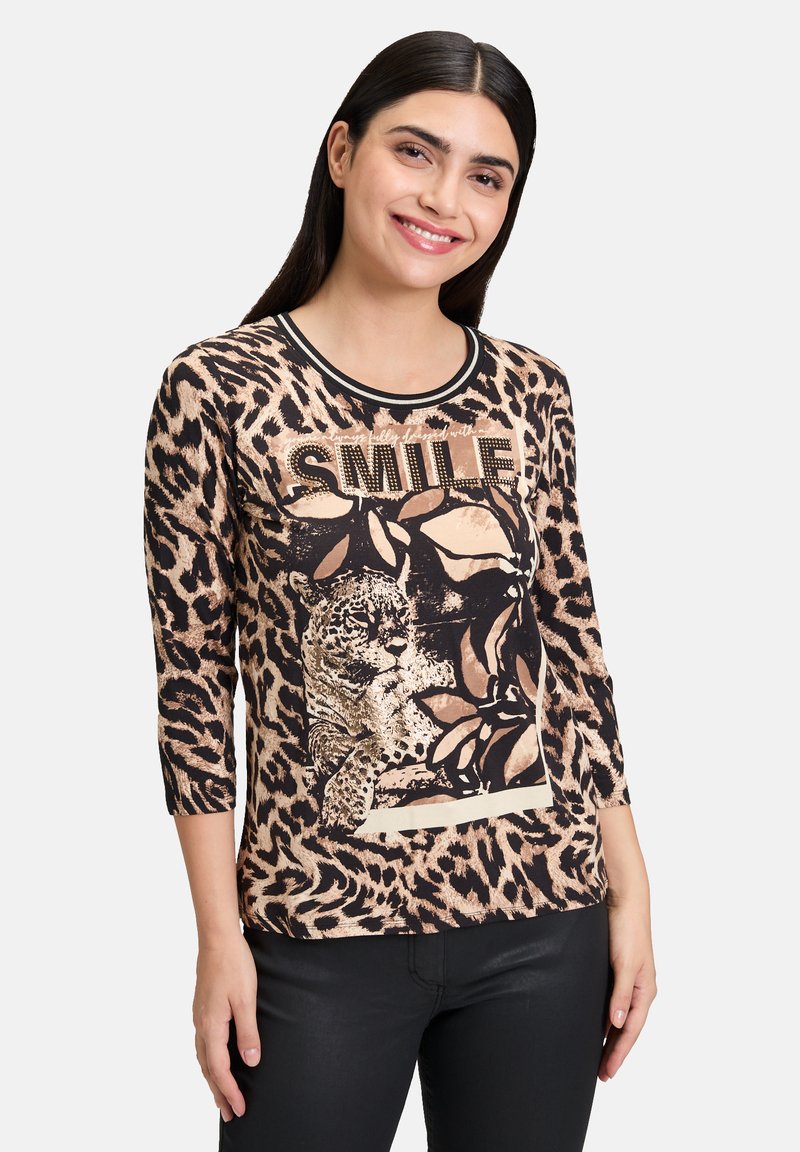 Haut à manches longues imprimé léopard avec des bordures noires, affichant un graphique de léopard et le mot "SMILE" en clous dorés, en tissu léger.