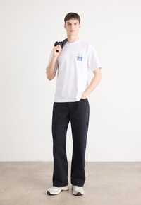 Hvit T-skjorte med en blåmønstret lomme, sammen med mørkeblå vide bukser og hvite sneakers. Modellen står med en hånd i lommen.
