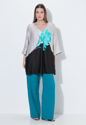 Kvinde iført en løs tunika med 3/4-ærmer med stort blåt blomstermotiv og teksturerede turkise bukser med vide ben, stående mod hvid baggrund.