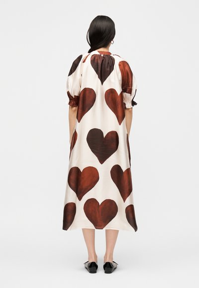 Γυναίκα με την πλάτη, φορώντας ένα λευκό midi φόρεμα με μεγάλα καφέ σχέδια καρδιάς, φουσκωτά μανίκια και μαύρα επίπεδα παπούτσια.