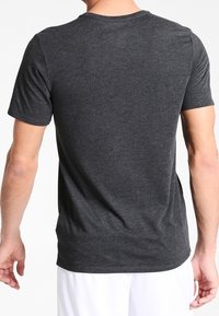 Mężczyzna w gładkiej, ciemnoszarej koszulce z krótkim rękawem, pokazany od tyłu, w połączeniu z białymi spodniami, na białym tle.