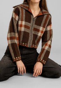 Donna seduta con le gambe incrociate, indossando una giacca zip-up a quadri marrone, arancione e beige e pantaloni neri, su uno sfondo semplice.