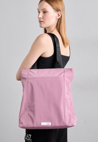 Ung kvinne med rett, rødt hår bærer en stor rosa tote bag med svarte EA7 Emporio Armani-stropper over skulderen.