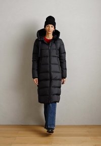 Vrouw gekleed in een lange zwarte gewatteerde jas met capuchon, blauwe jeans, een zwarte muts en zwarte laarzen tegen een effen lichte muur.