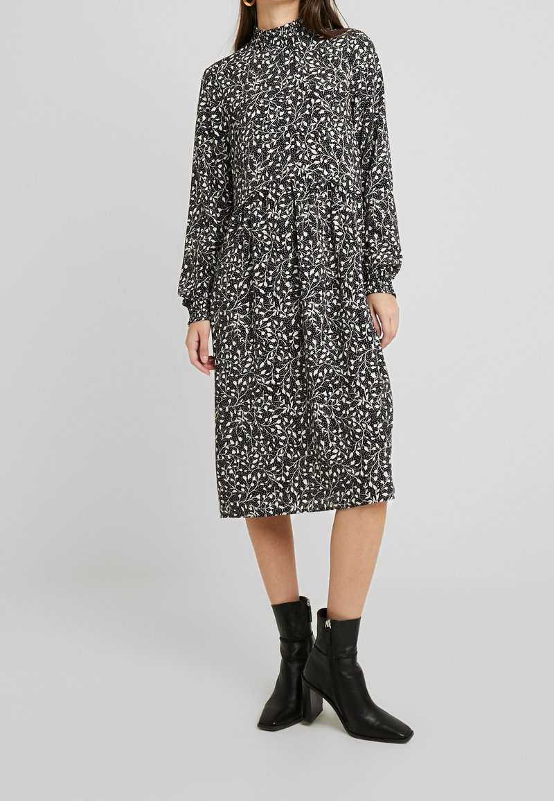 Femme portant une robe midi à manches longues noire et blanche à motif floral avec un col haut, associée à des bottines noires à talons.