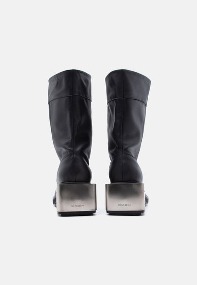 Ζευγάρι μαύρων δερμάτινων μποτών μέσου μήκους που φτάνουν μέχρι τη μέση της γάμπας με μεταλλικές τακούνια τύπου block, απεικονιζόμενα από πίσω πάνω σε λευκό φόντο.