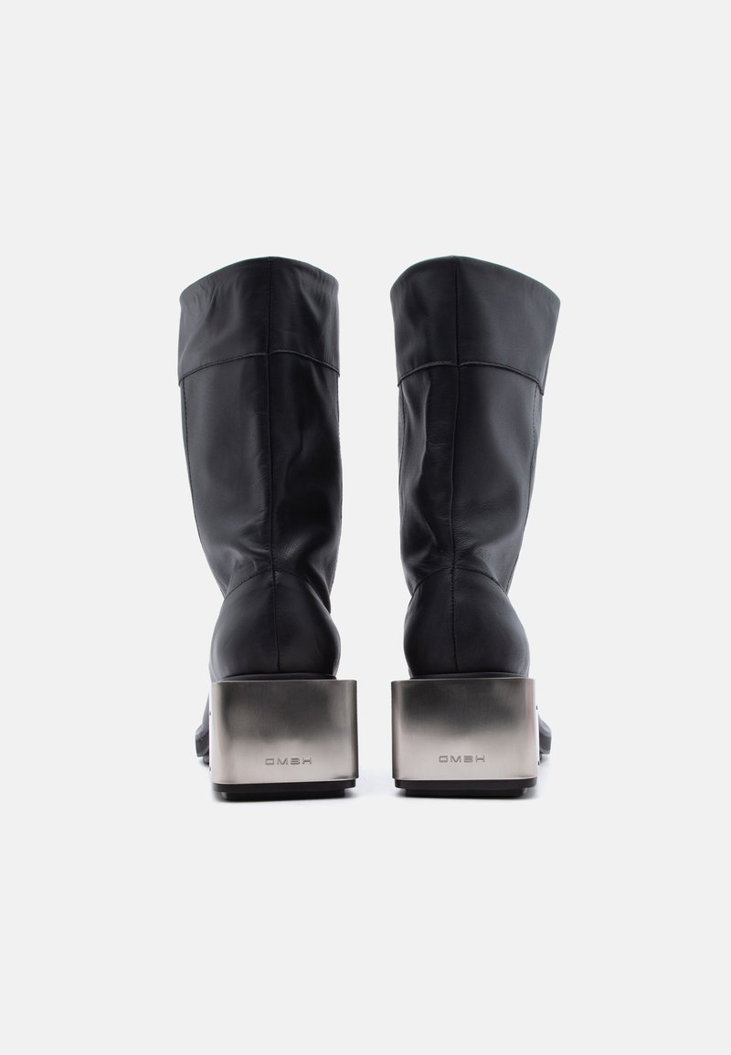 Coppia di stivali neri in pelle a metà polpaccio con tacchi a blocco metallici mostrati di spalle su sfondo bianco.