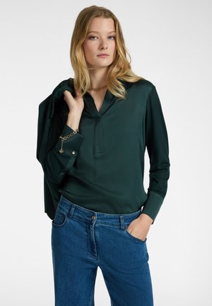 Blusa verde scuro a maniche lunghe con colletto e patta frontale. Abbinata a jeans blu, che mettono in mostra una texture liscia e morbida.