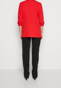 Röd kavaj med trekvartsärmar, strukturerad design och rundad krage, i kombination med svarta byxor och klackar. En enkel och skräddarsydd look.