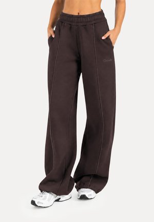 Mujer con pantalones deportivos negros holgados con bolsillos laterales y zapatillas blancas, de pie con las manos en los bolsillos frente a un fondo liso.