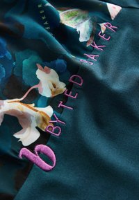 Dunkles Gewebe mit einem floralen Muster in Blau- und Cremetönen, mit pinkfarbenem besticktem Text "6 BY TED BAKER". Glatte Textur.