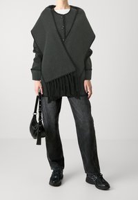 Poncho grigio scuro con frange, caratterizzato da cuciture decorative e bottoni. Abbinato a jeans neri larghi e sneaker, tiene in mano una borsa nera.
