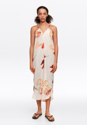 Kobieta stoi, mając na sobie kremową, bezrękawnikową sukienkę midi z pomarańczowymi kwiatowymi i liściastymi wzorami oraz cienkimi ramiączkami, w zestawieniu z czarnymi sandałami.