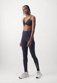 Black sports bra with a twist front detail, paired with navy high-waisted leggings featuring a logo print. Gray athletic shoes complete the outfit.