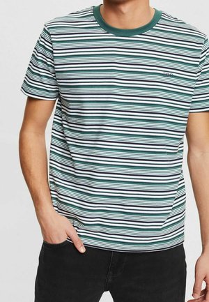 Mand iført en kortærmet t-shirt med grønne, hvide og sorte vandrette striber og sorte bukser, den ene hånd i lommen.