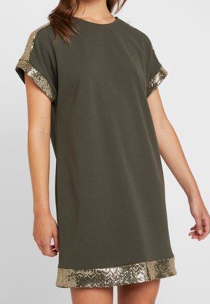 Femme portant une robe courte à manches courtes vert olive avec bordure en sequins dorés aux manches et à l'ourlet, debout devant un fond clair uni.