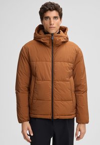 Giacca puffa marrone con cappuccio, dotata di chiusura con zip, polsini elastici e texture trapuntata. Indossata sopra uno strato base nero.
