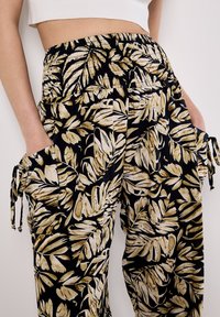 Pantalons noirs avec un motif doré et crème de feuilles, dotés d'une taille élastique et de poches latérales avec des cordons de serrage.
