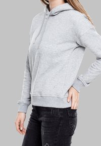 Mujer vistiendo una sudadera ligera gris y jeans negros desgastados, con una mano en el bolsillo trasero, contra un fondo gris liso.