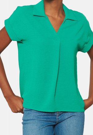 Persoon draagt een felgroen poloshirt met korte mouwen en opgerolde manchetten, en een blauwe spijkerbroek met handen in de zakken.