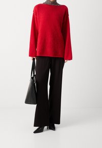 Persona che indossa un maglione rosso di pelliccia sopra un dolcevita a righe bianche e nere, pantaloni neri a gamba larga, tacchi a punta neri, e tiene una borsa a tracolla nera.