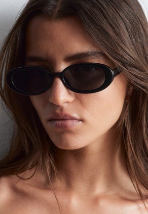 Donna con capelli castani lunghi che indossa piccoli occhiali da sole neri ovali, rivolta leggermente a sinistra con espressione neutra e spalle scoperte visibili.