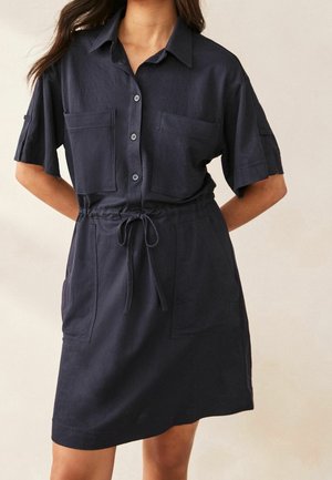 Woman wearing a short-sleeve, button-up navy dress with a collar, front pockets, and a drawstring waist against a plain background.