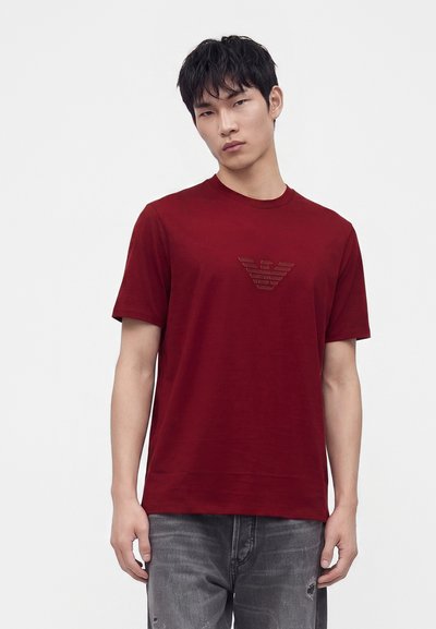 Νέος άνδρας φοράει κόκκινο T-shirt με σκούρο λογότυπο στο στήθος και ξεθωριασμένα γκρίζα τζιν, στέκεται μπροστά σε απλό ανοιχτό φόντο.