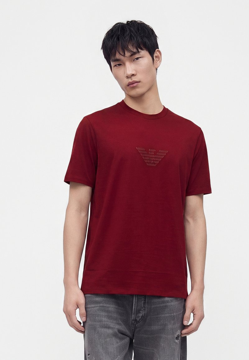 Νέος άνδρας φοράει κόκκινο T-shirt με σκούρο λογότυπο στο στήθος και ξεθωριασμένα γκρίζα τζιν, στέκεται μπροστά σε απλό ανοιχτό φόντο.