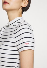 Top de rayas en blanco y marino con canalé, con cuello alto, que presenta mangas cortas y el logo rojo "HUGO" en la manga izquierda.