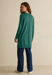 Långärmad grön cardigan med ribbad nederkant och ärmslut, som har en avslappnad passform och detalj med söm bak, kombinerad med lösa marinblå jeanstights och sneakers.