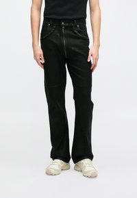 Crne traperice s prednjim zatvaračem na patentni zatvarač, kopčanjem na dugme i sirovim rubovima. Imaju ravni kroj i suptilne detalje šivanja.