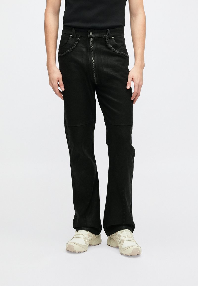 Crne traperice s prednjim zatvaračem na patentni zatvarač, kopčanjem na dugme i sirovim rubovima. Imaju ravni kroj i suptilne detalje šivanja.