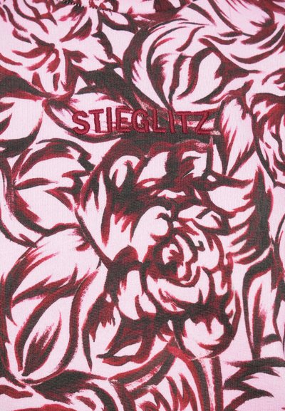 Tejido con patrón de rosas en tonos de rojo y rosa, con la palabra bordada "STIEGLITZ" en rojo sobre un fondo texturizado.